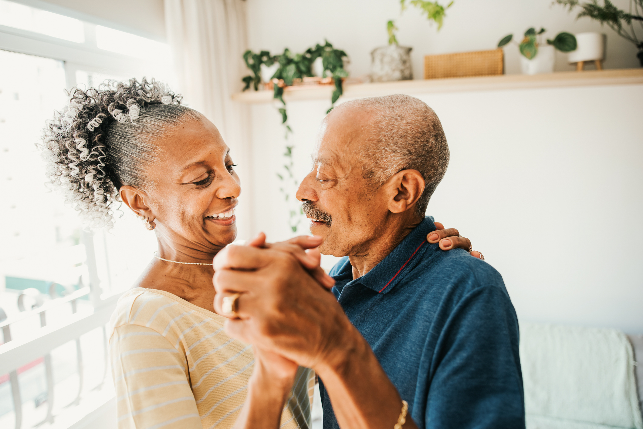A couple dances together in their kitchen in afternoon light. They have medium to dark skin tones and gray hair and smile sweetly at each other.